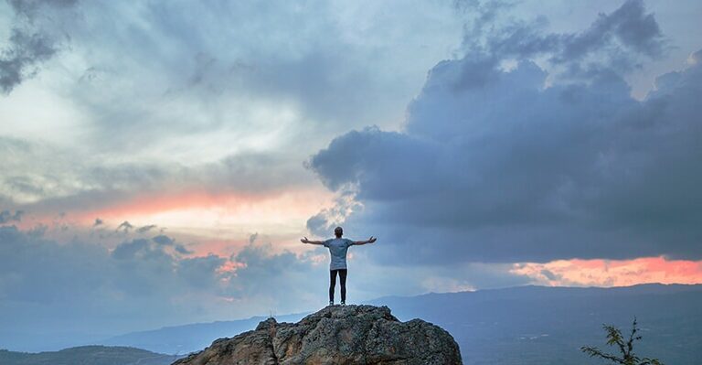 Person standing with arms outstretched on a rocky hill beneath a blue evening sky